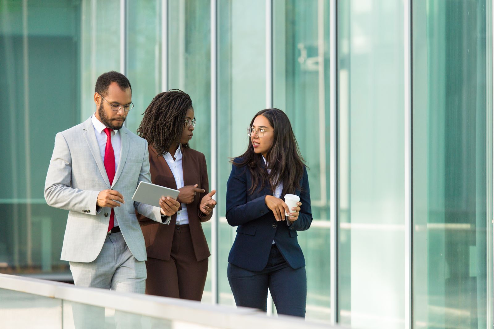 Excited business colleagues going along outdoor office glass wall. Business man and women walking outside, using tablet, drinking coffee, talking. Business group concept