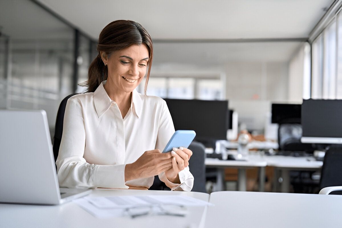 Smiling businesswoman using a smartphone while seated at a modern office desk with a laptop and documents – Neutrinos