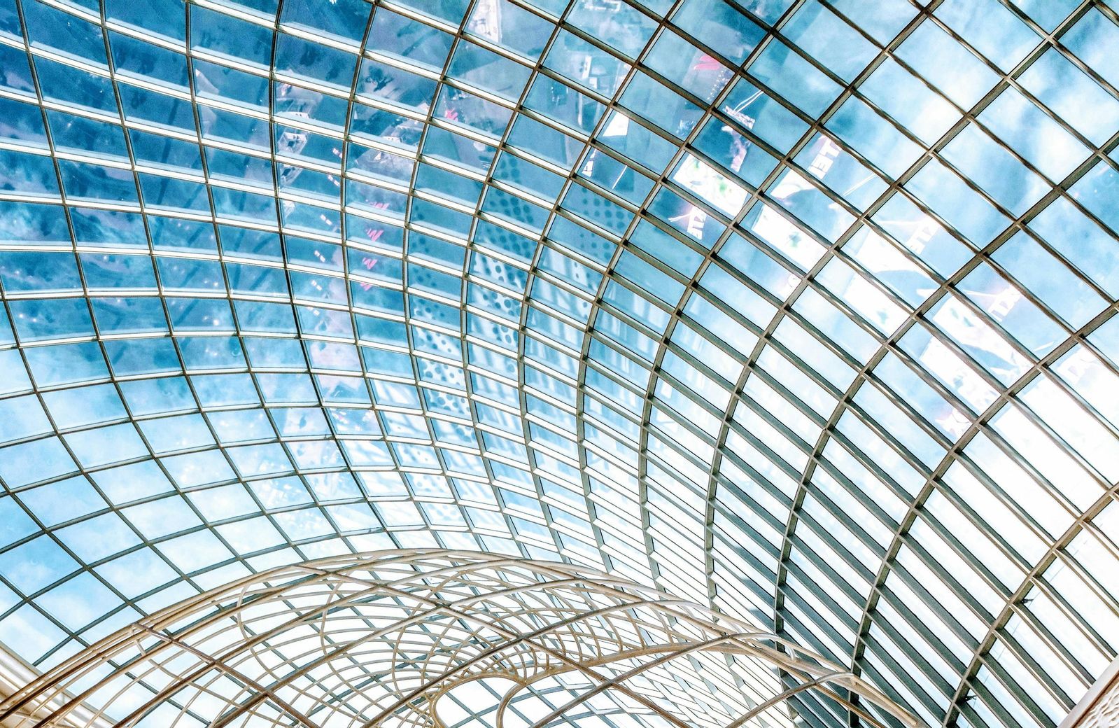 Modern glass ceiling architecture with a curved grid design against a blue sky – Neutrinos.