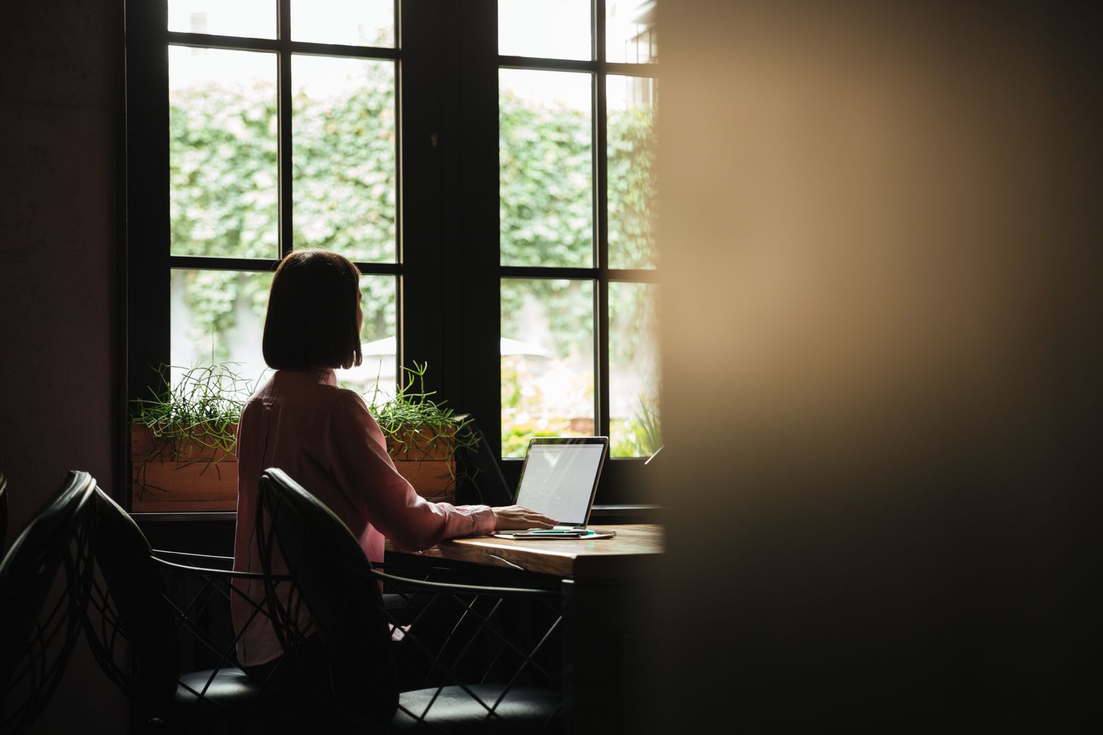 Back view of brunette woman sitting by the table near window and using laptop computer
