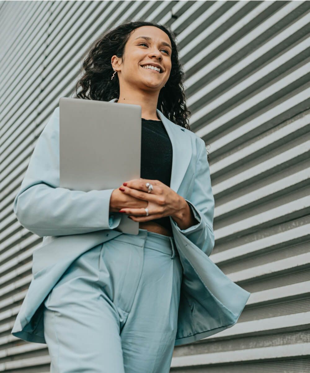 Confident businesswoman in a light blue suit walking outdoors with a laptop in hand, smiling.-Neutrinos
